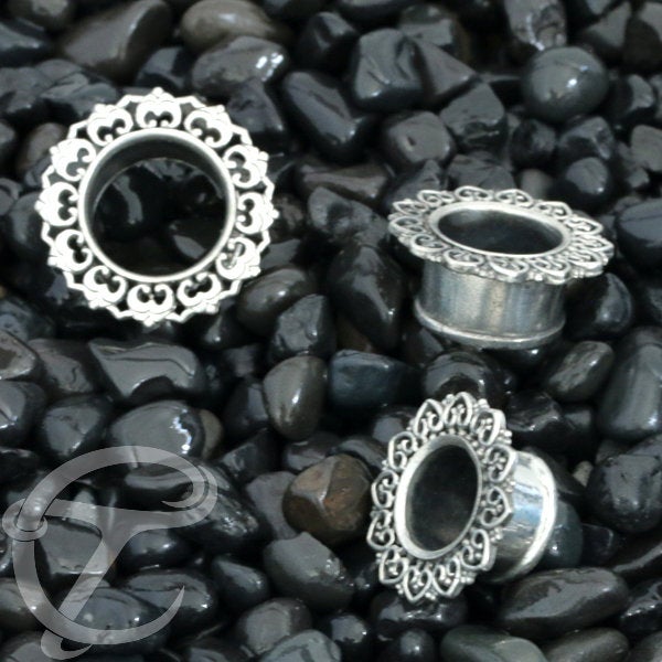 Three ornate silver ear gauges from Kaya Movement's ONAS Sacred Lotus Mandala Round Tunnels collection are displayed on shiny black pebbles. The gauges, reminiscent of tribal plugs, have a circular shape with decorative edges. One gauge stands upright, while the other two lie on their sides, showcasing the craftsmanship of these finely detailed jewelry pieces available in sizes ranging from 3mm to 18mm.